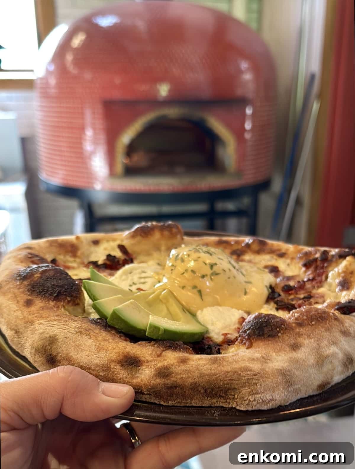 A hand holds a plate with a wood-fired pizza topped with lime wedges, sauce, and herbs; a red pizza oven is visible in the background.