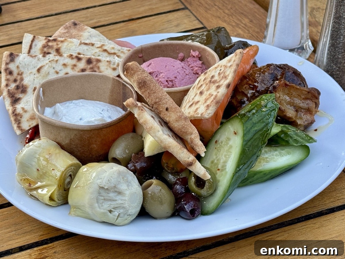 A white plate with pita bread, grape leaves, cucumber slices, olives, artichoke hearts, dips, and other Mediterranean appetizers on a wooden table.