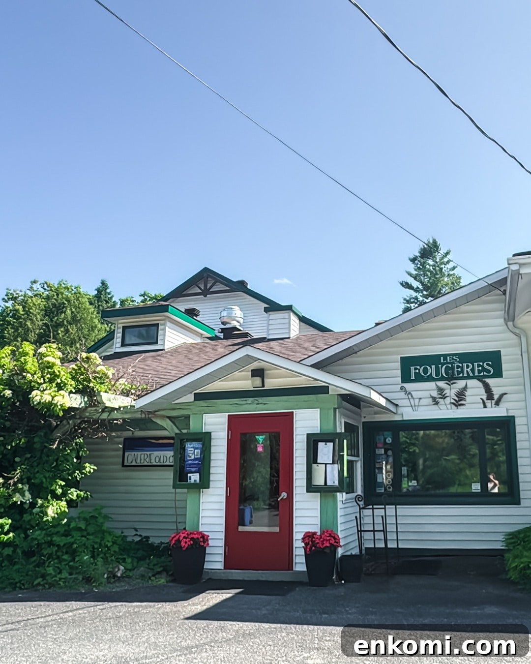 A small white building with green trim and a red door houses a restaurant named "Les Fougères," surrounded by greenery under a clear blue sky.