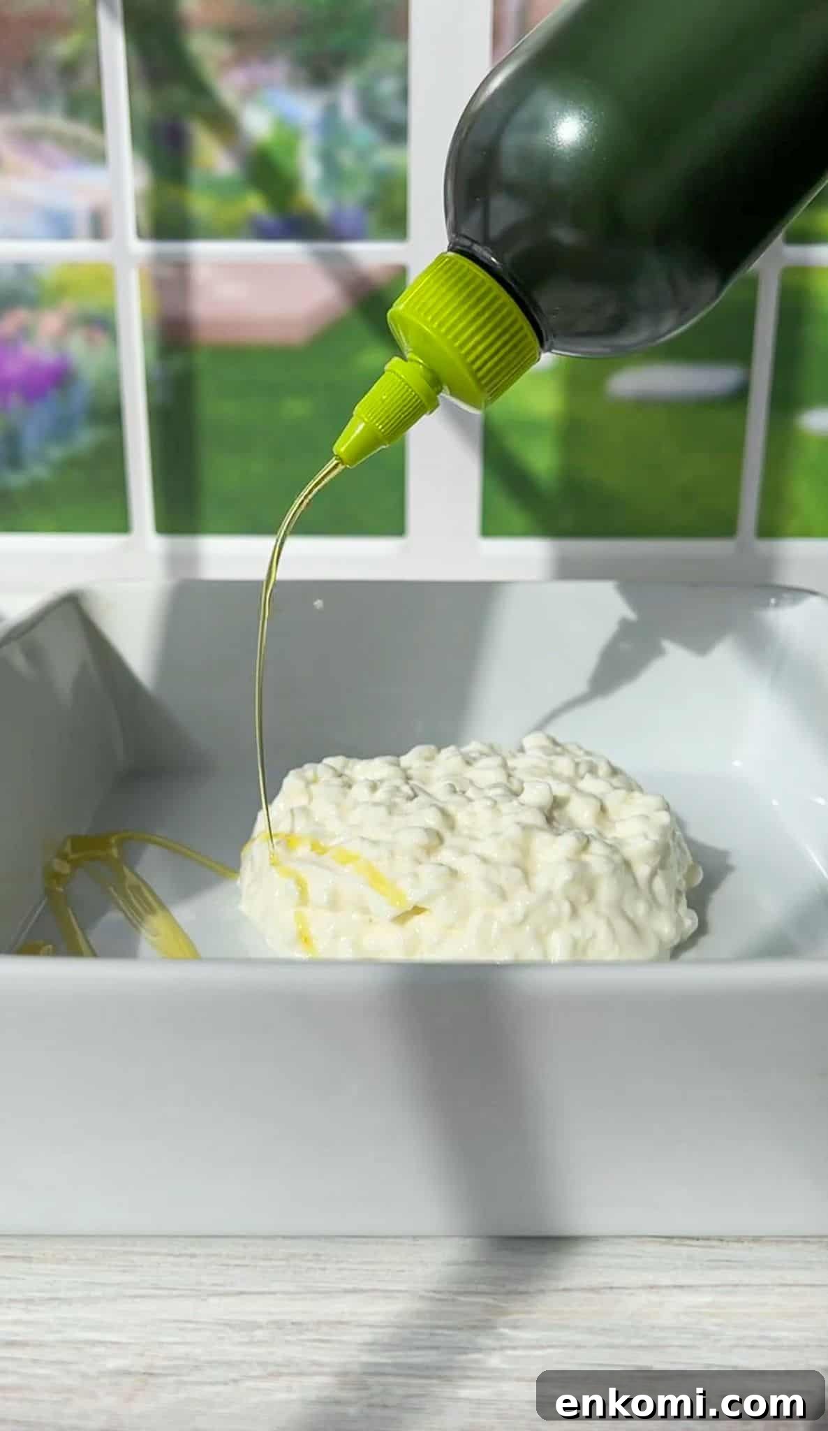 Cottage Cheese Scrambled Eggs Recipe for Creamy Protein-Packed Breakfast 3 A black bottle with a green nozzle pours oil over a round piece of burrata cheese in a white square dish. The background features blurred garden scenery visible through a window.