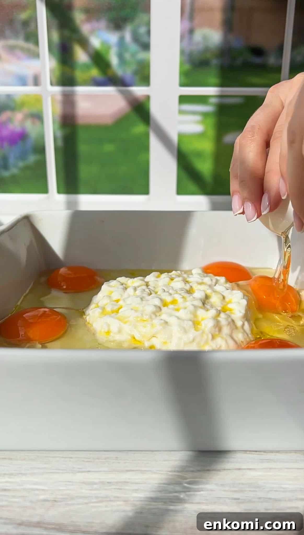 Cottage Cheese Scrambled Eggs Recipe for Creamy Protein-Packed Breakfast 4 A hand cracking an egg into a white baking dish containing several eggs and a pile of cottage cheese. The background shows a window with a view of a garden.