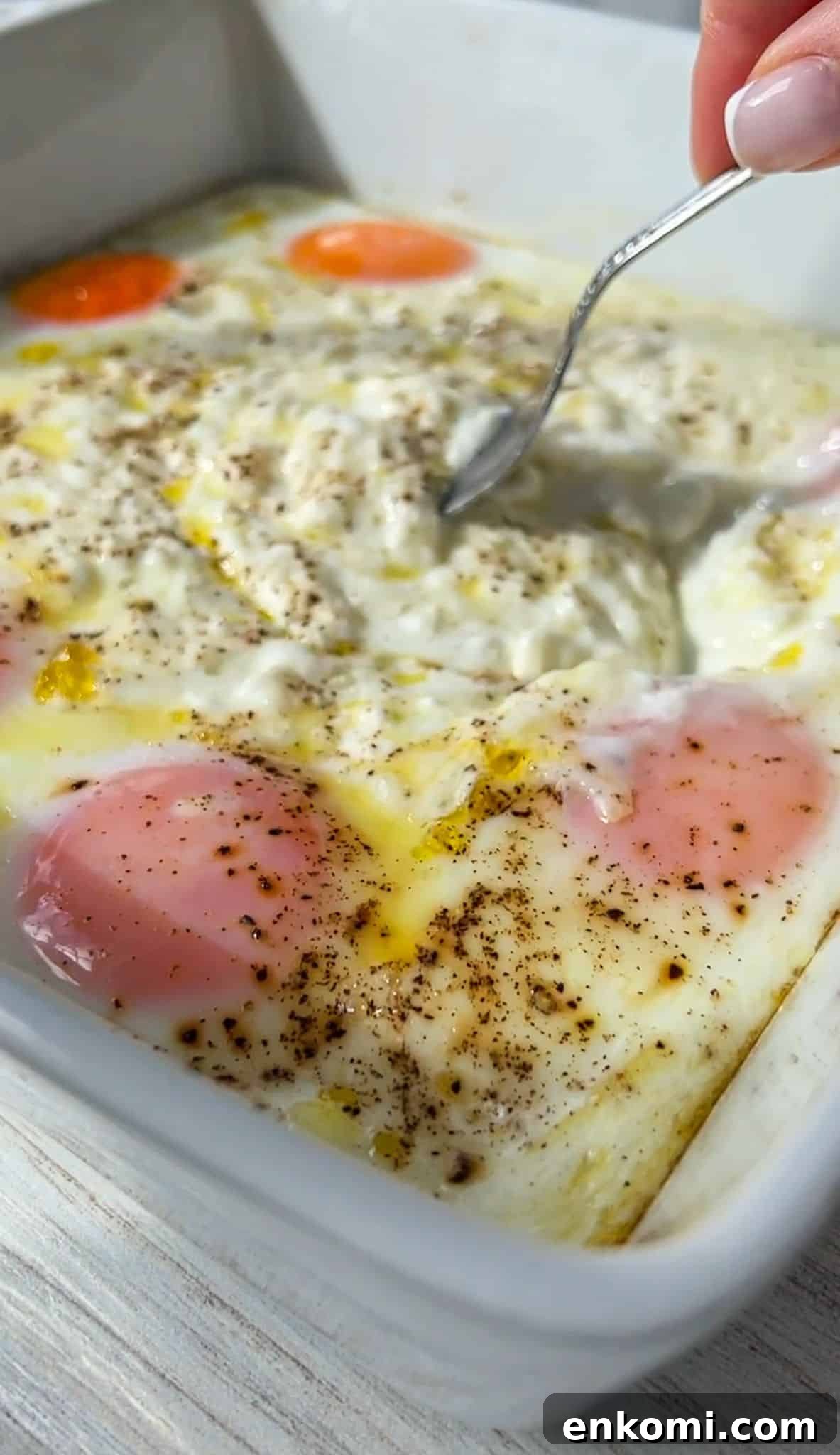 Cottage Cheese Scrambled Eggs Recipe for Creamy Protein-Packed Breakfast 5 A spoon is stirring a dish with poached eggs seasoned with pepper in a white baking dish. The eggs are nestled in a creamy sauce.