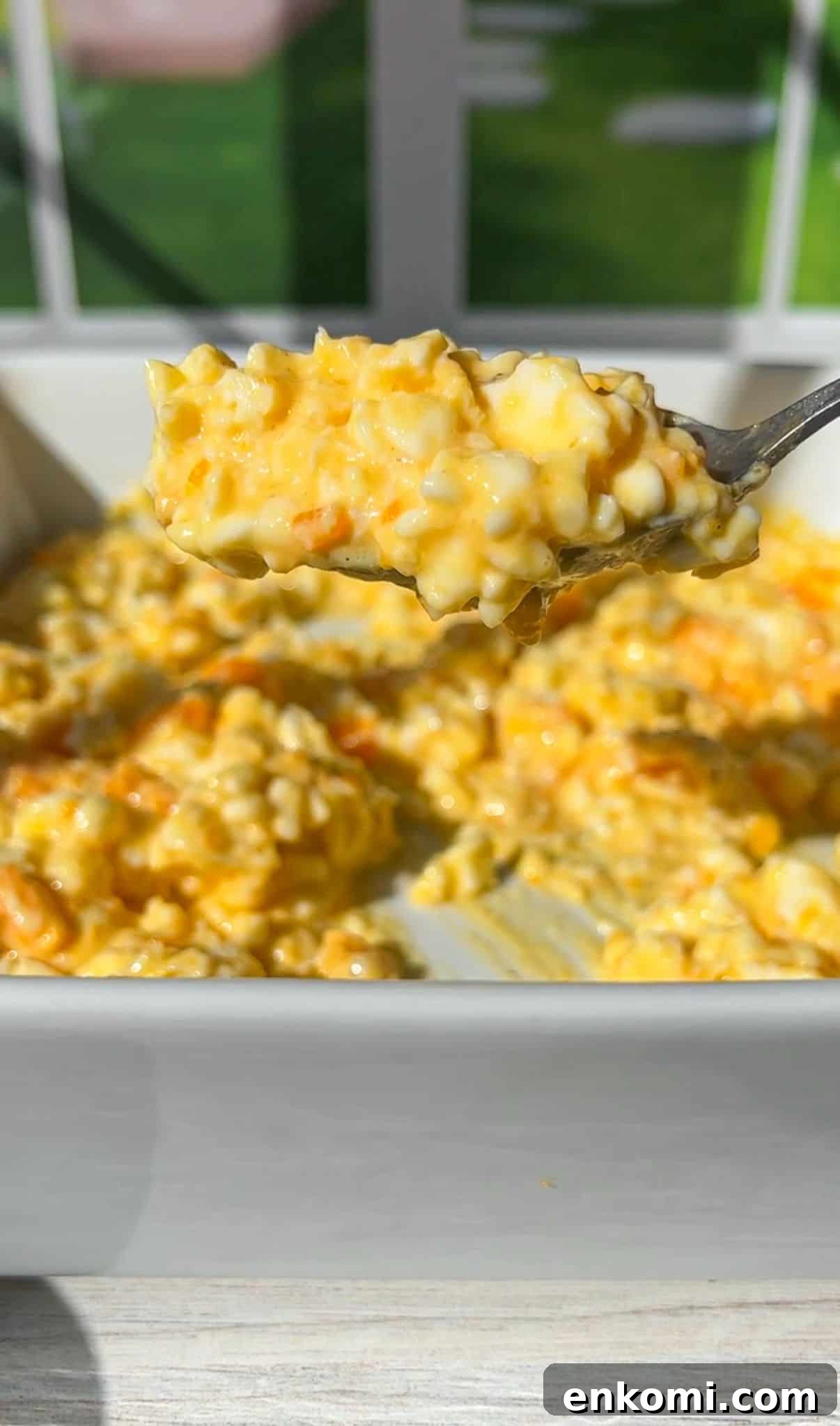 Cottage Cheese Scrambled Eggs Recipe for Creamy Protein-Packed Breakfast 6 A spoonful of cottage cheese eggs is held above a white baking dish. The dish is filled with creamy, golden casserole, with visible chunks of potatoes and melted cheese. A sunlit window is blurred in the background.