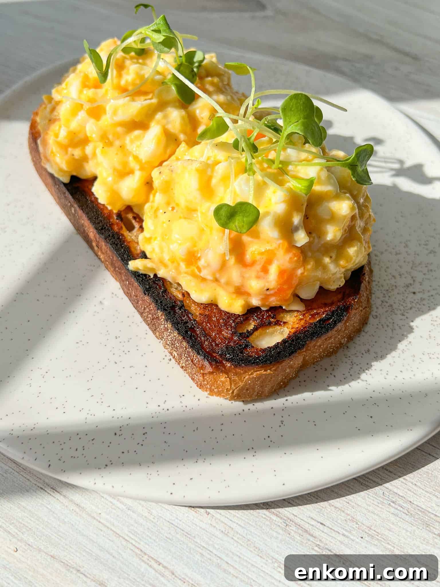 Cottage Cheese Scrambled Eggs Recipe for Creamy Protein-Packed Breakfast 7 The perfect cottage cheese eggs. Open-faced sandwich with scrambled eggs on toasted bread, garnished with microgreens. The sandwich is on a white plate, and sunlight is casting shadows, enhancing the texture of the dish.