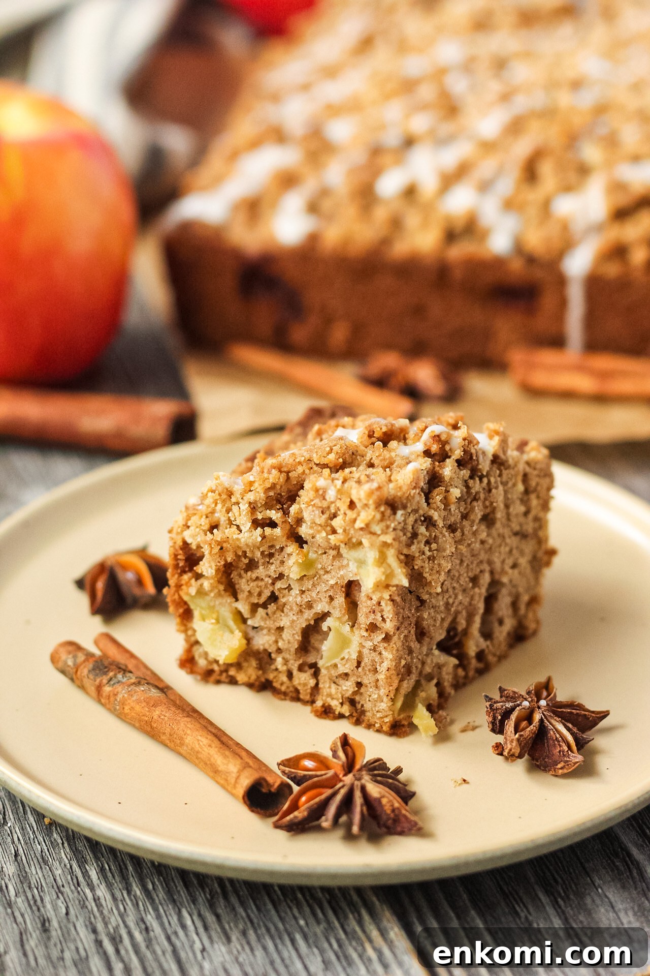 side shot of slice of apple crumb cake on a brown plate with apples and more cake in the background.