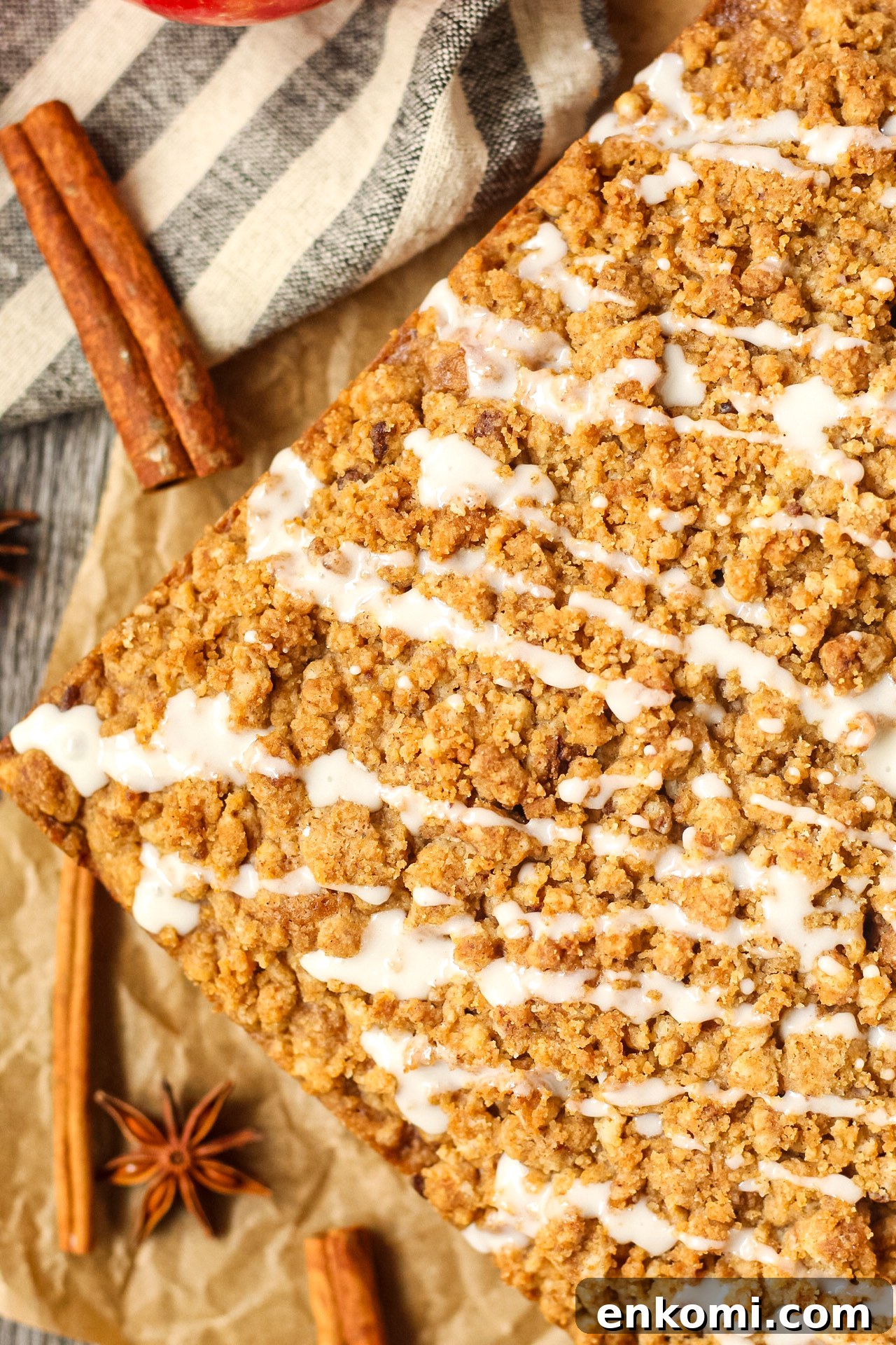 close up overhead shot of spiced apple crumb cake on parchment paper on top of wooden surface with apples and cinnamon sticks in the perimeter of the shot.