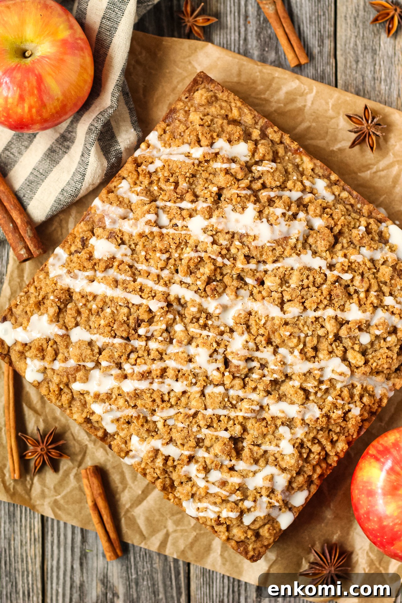 overhead shot of spiced apple crumb cake on parchment paper on top of wooden surface with apples and cinnamon sticks in the perimeter of the shot. 