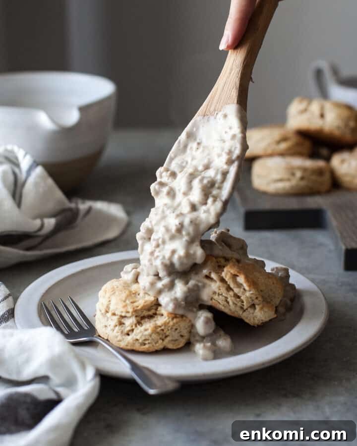 healthy biscuits and gravy on a white plate with a fork on the plate and a woman putting more gravy on the biscuits with a wooden spoon, all on a gray table
