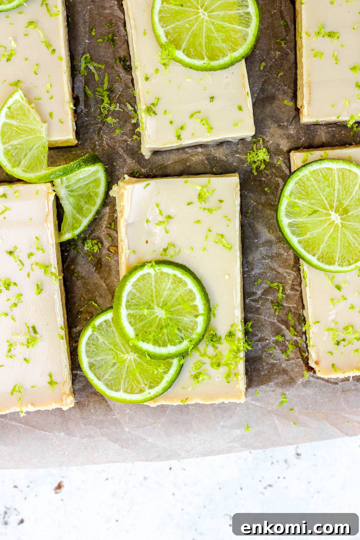 Key lime pie bars with fresh lime slices and lime zest on brown parchment paper.