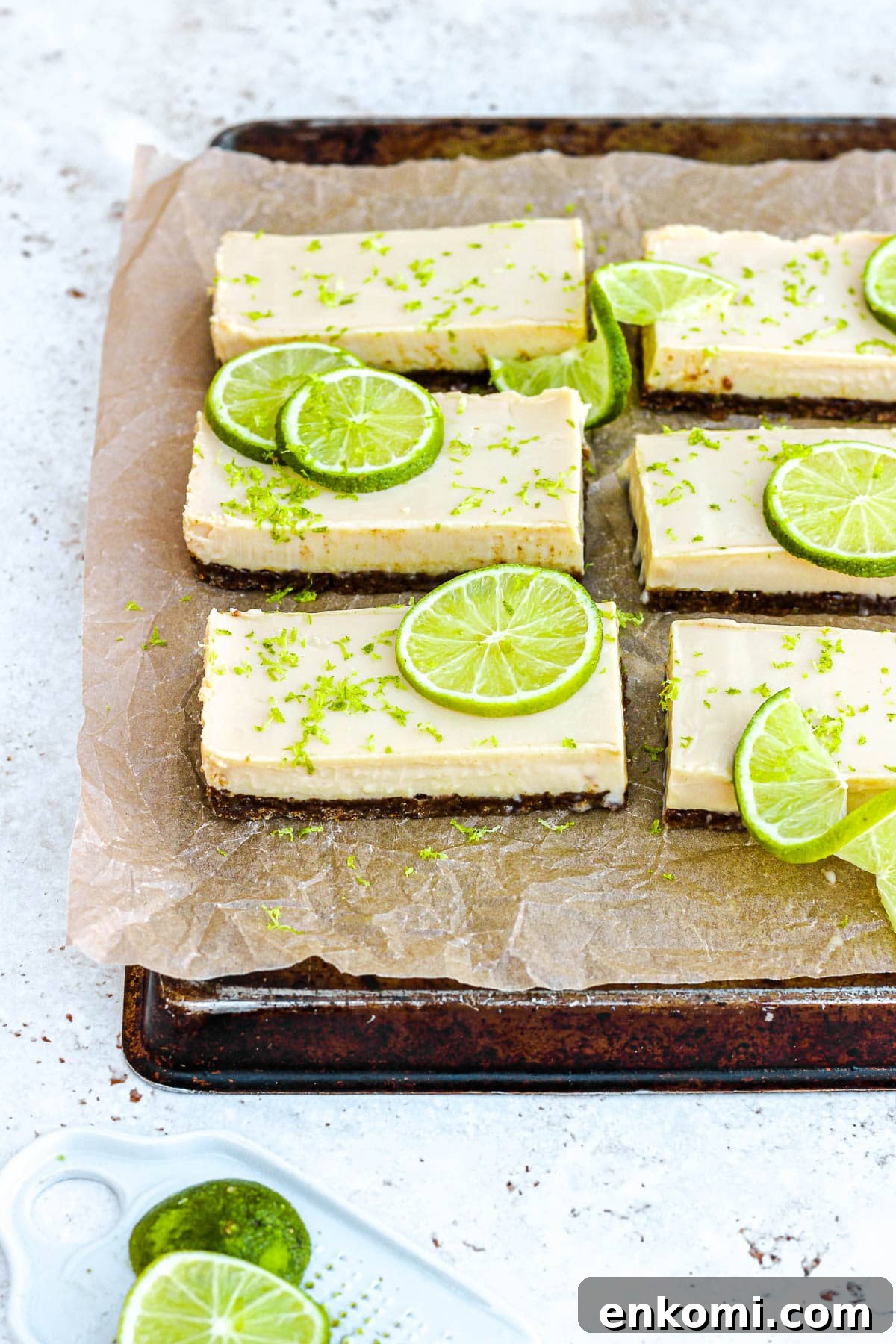 Key lime pie bars with fresh lime slices and lime zest on brown parchment paper.