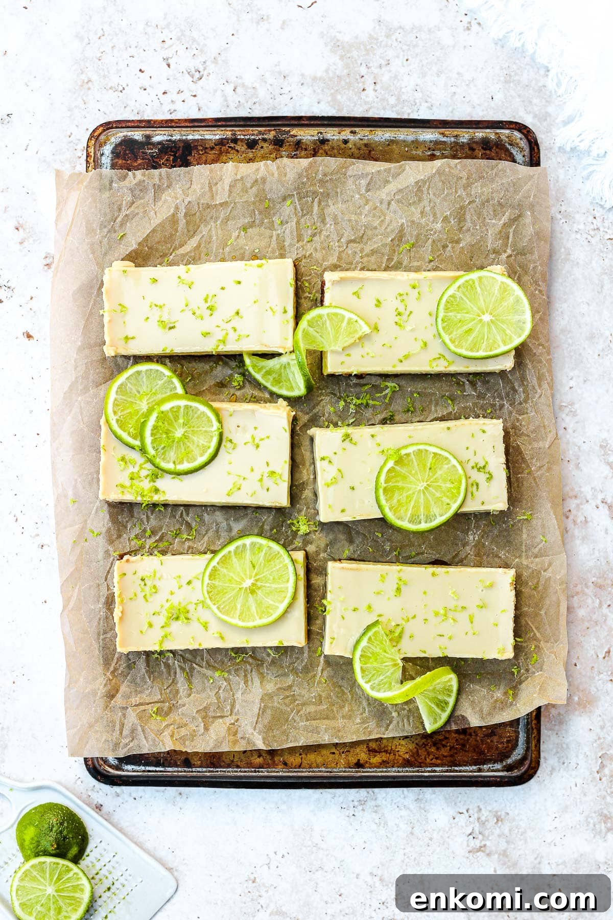 Key lime pie bars with fresh lime slices and lime zest on brown parchment paper.