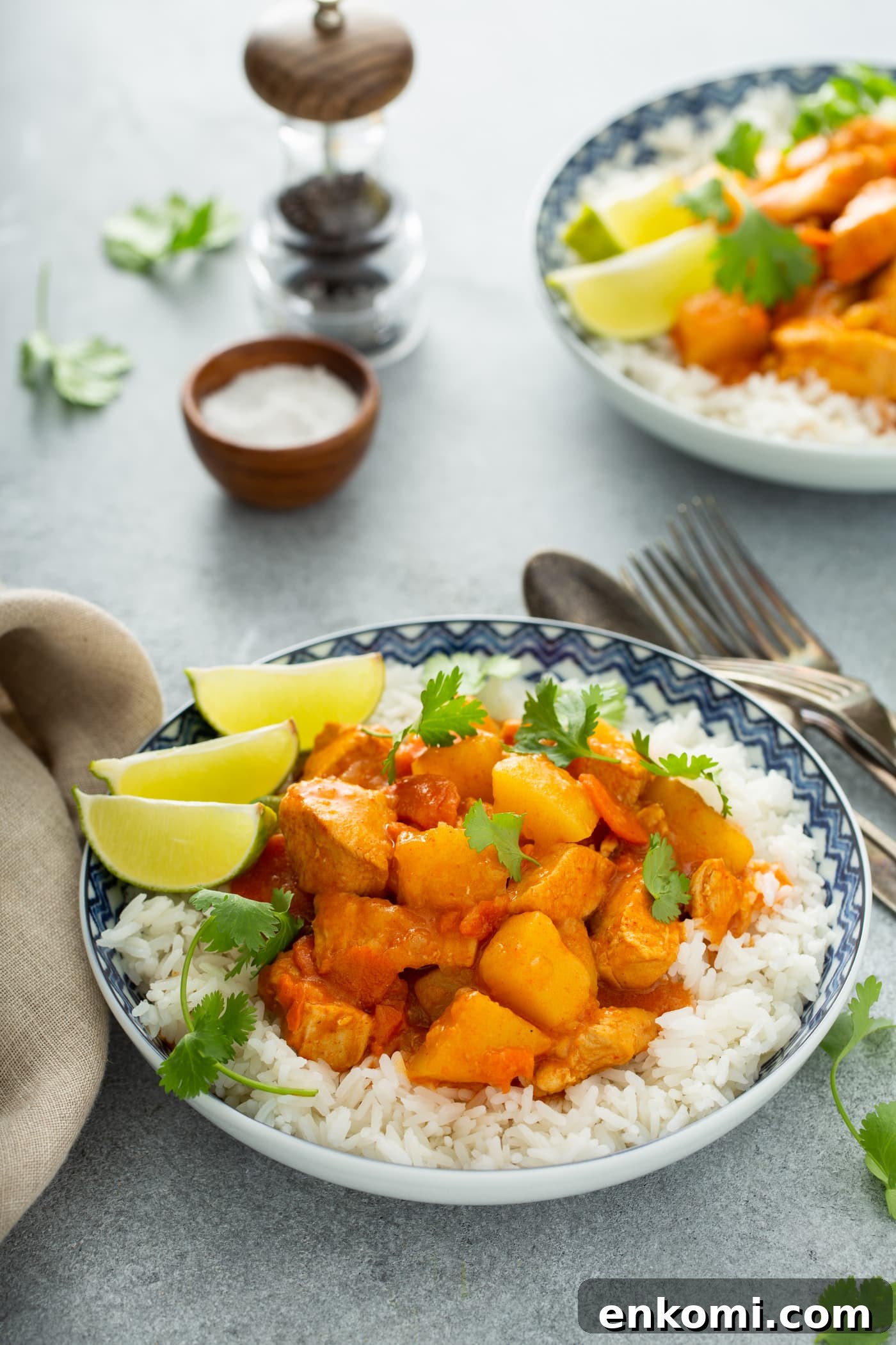 A bowl of white rice with chicken curry on top. There are chunks of chicken, potatoes and carrots in a curry sauce. Three lime wedges are on the edge of the bowl and fresh parsley is sprinkled on top. There are forks and spoons next to the bowl. In the background is a container of salt and pepper and another serving of chicken curry.