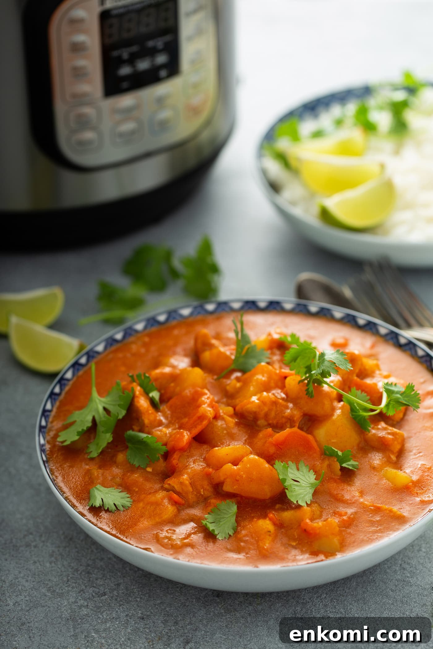 A bowl of chicken curry with chunks of chicken, potatoes and carrots in a curry sauce. Fresh parsley leaves are sprinkled on top and there is an instant pot, and lime wedges in the background.