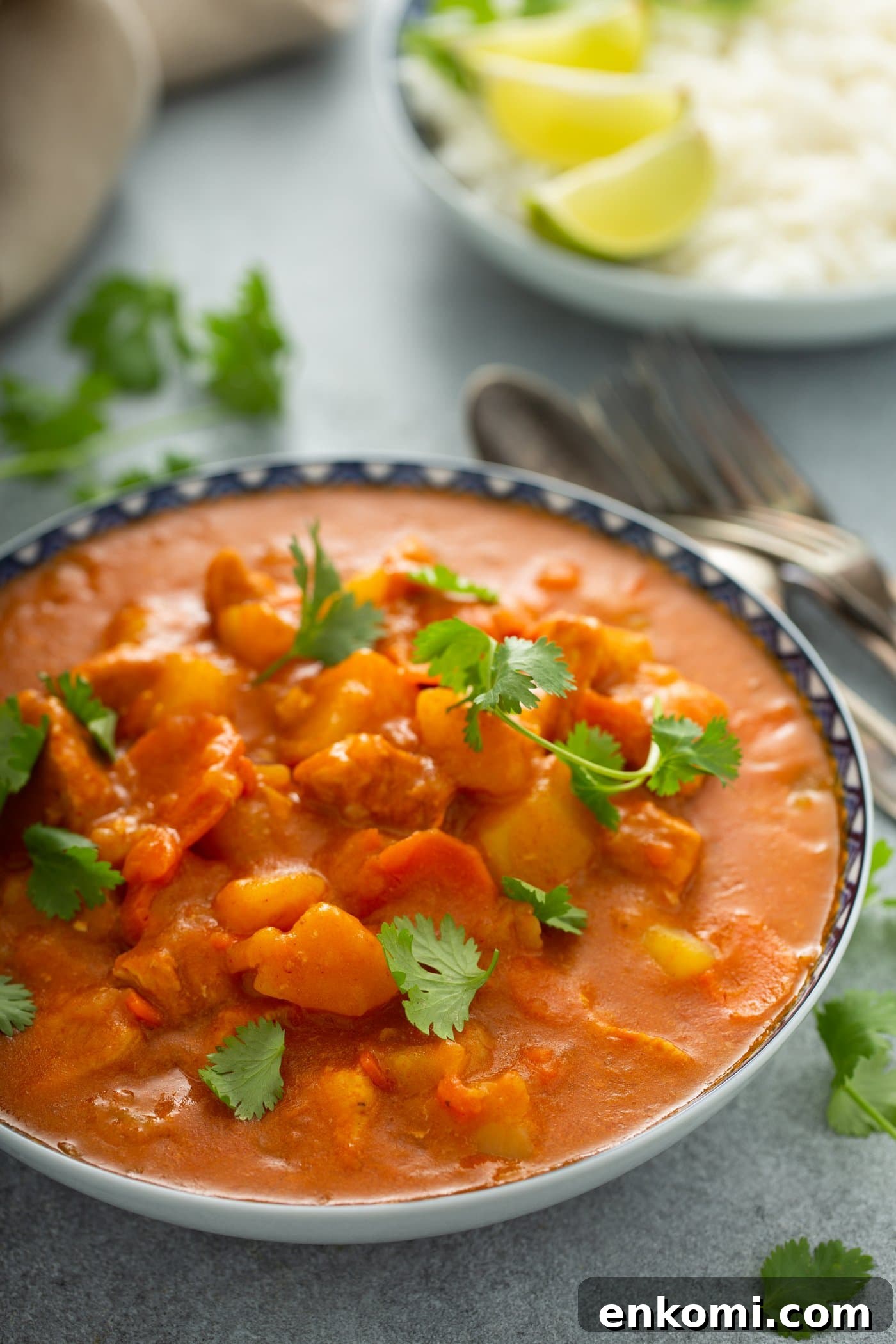 A bowl of chicken curry with chunks of chicken and potatoes and carrots in a curry sauce. Parsley leaves are scattered on top and on the table next to the curry. A fork and spoon and a plate with lime wedges are in the background.