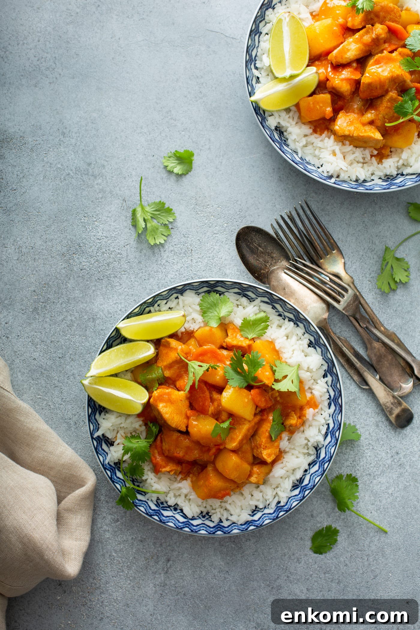A bowl of rice topped with chicken curry. There are lime wedges on the edge of the bowl and forks and spoons next to the bowl. Another bowl of curry is in the background.