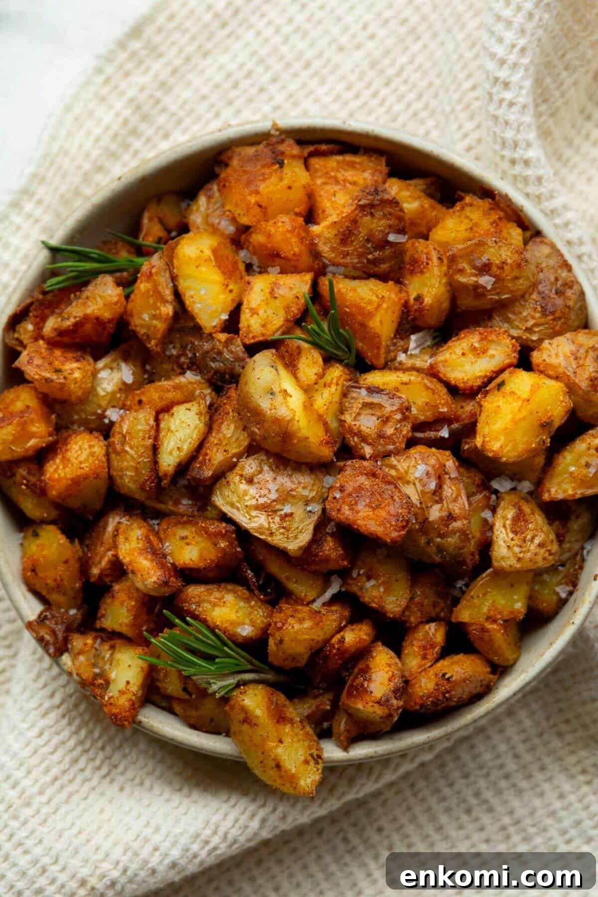 Bowl of crispy oven roasted potatoes with golden brown edges, garnished with fresh rosemary sprigs and flaky sea salt.