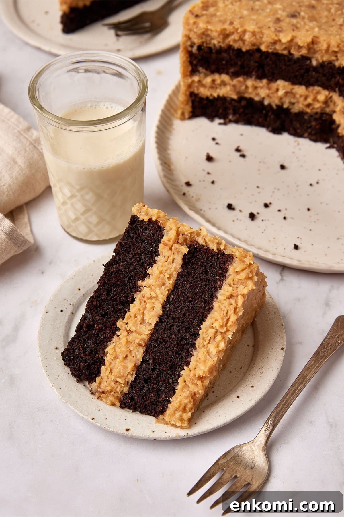 Keto German Chocolate Cake Recipe: Low-Carb Decadent Dessert 2 Slice of German Chocolate cake sits on a small white plate next to a fork. A glass of milk is sitting next to it and the cut cake is just visible in the background.