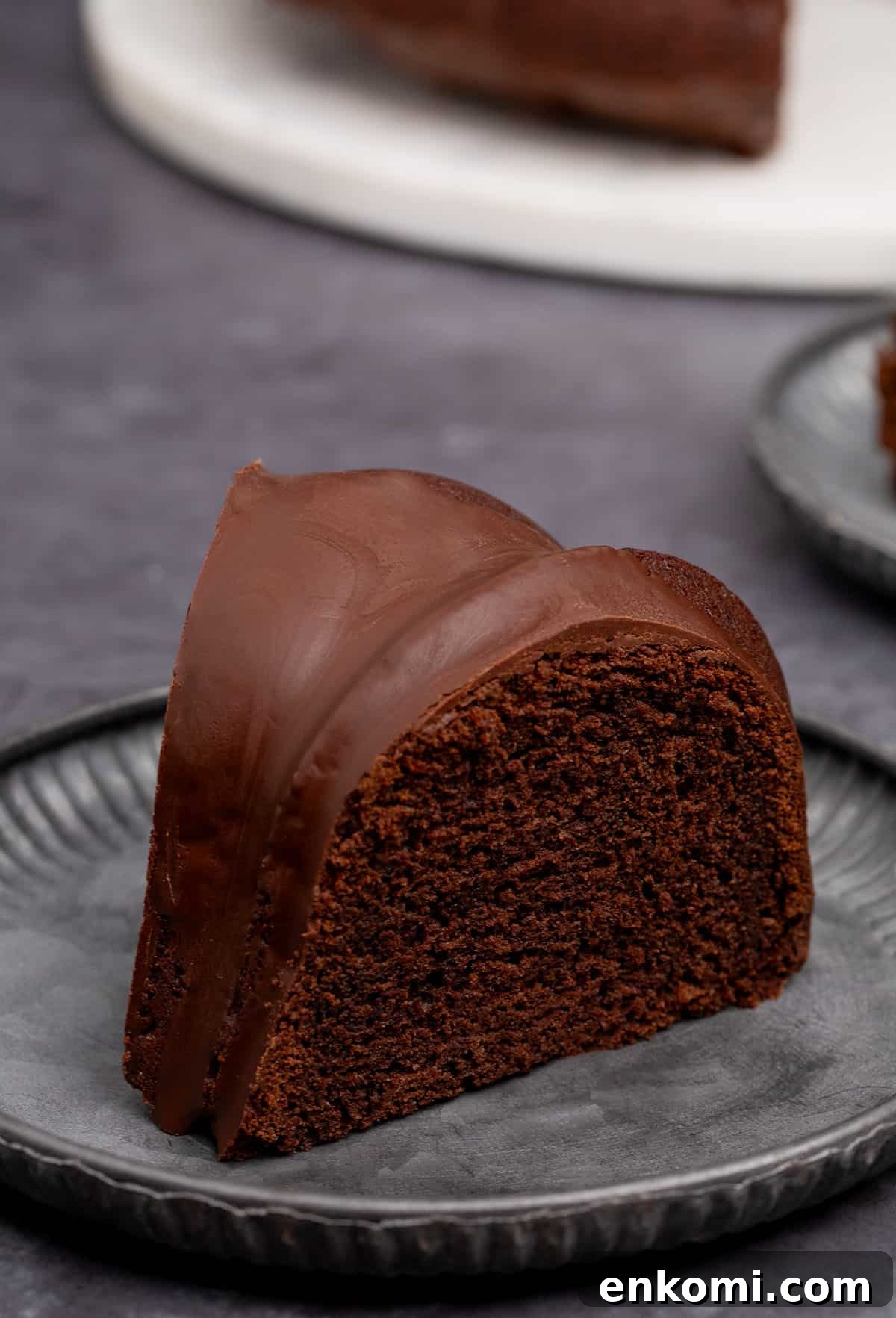 side view of slice of dark brown chocolate bundt cake on dark gray fluted cake plate with rest of cake on white cake plate in background