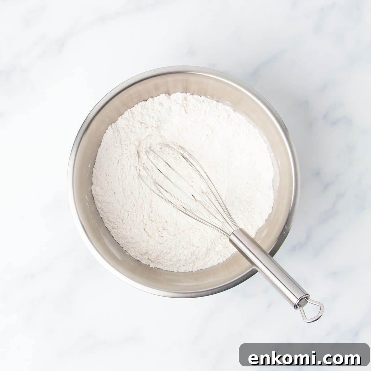 medium-size round metal mixing bowl with white flour powder and large wire whisk
