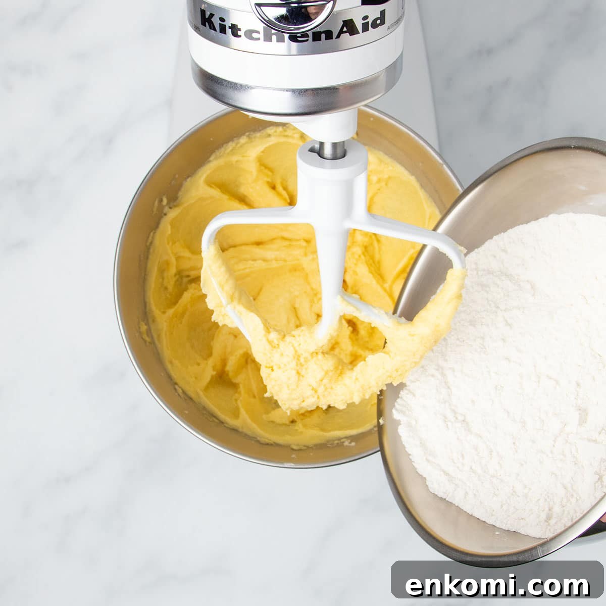 darker yellow mixture in stand mixer bowl with white powder contents of medium bowl being added to the mixing bowl