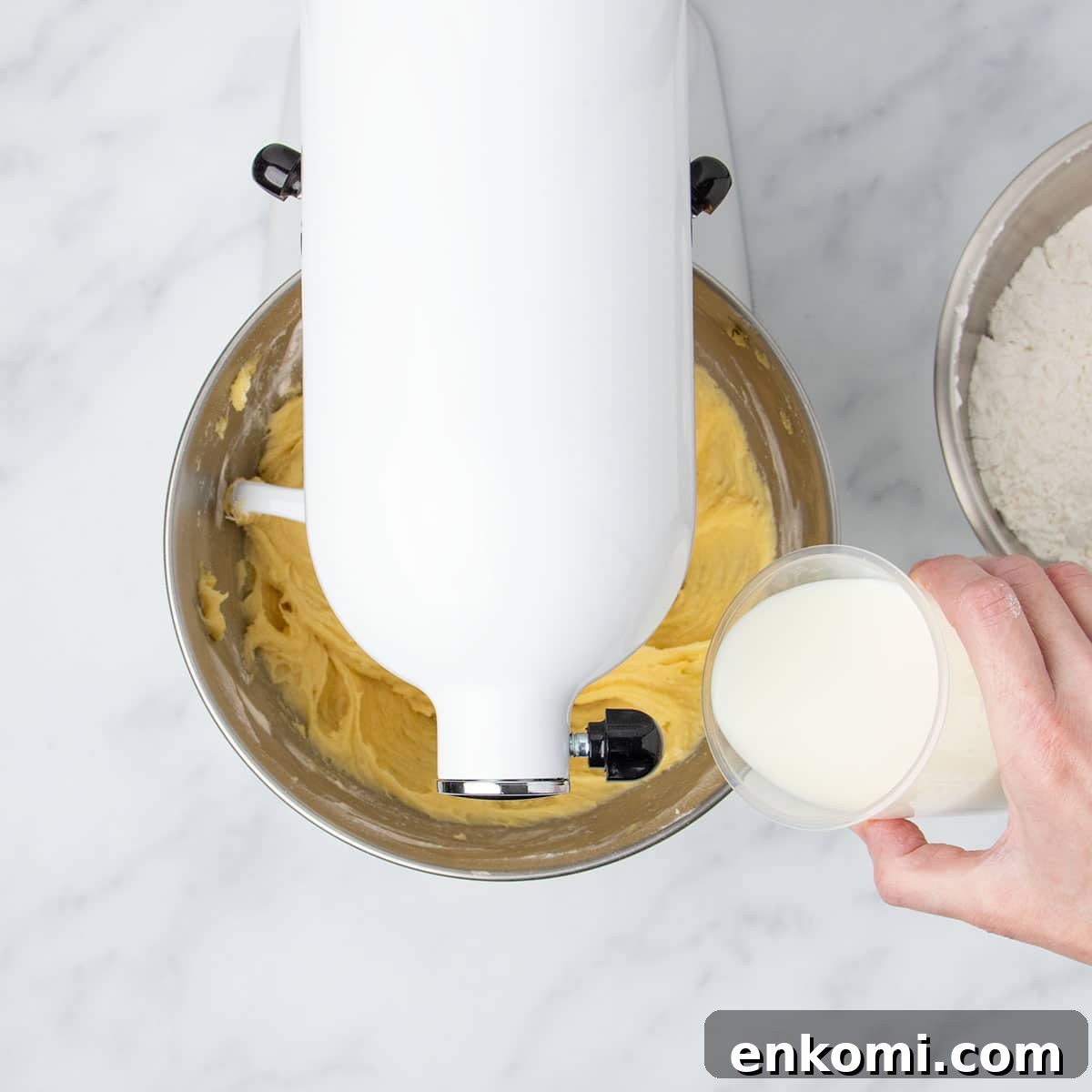 white stand mixer with head tilted down with hand tipping milk in plastic measuring cup into mixing bowl