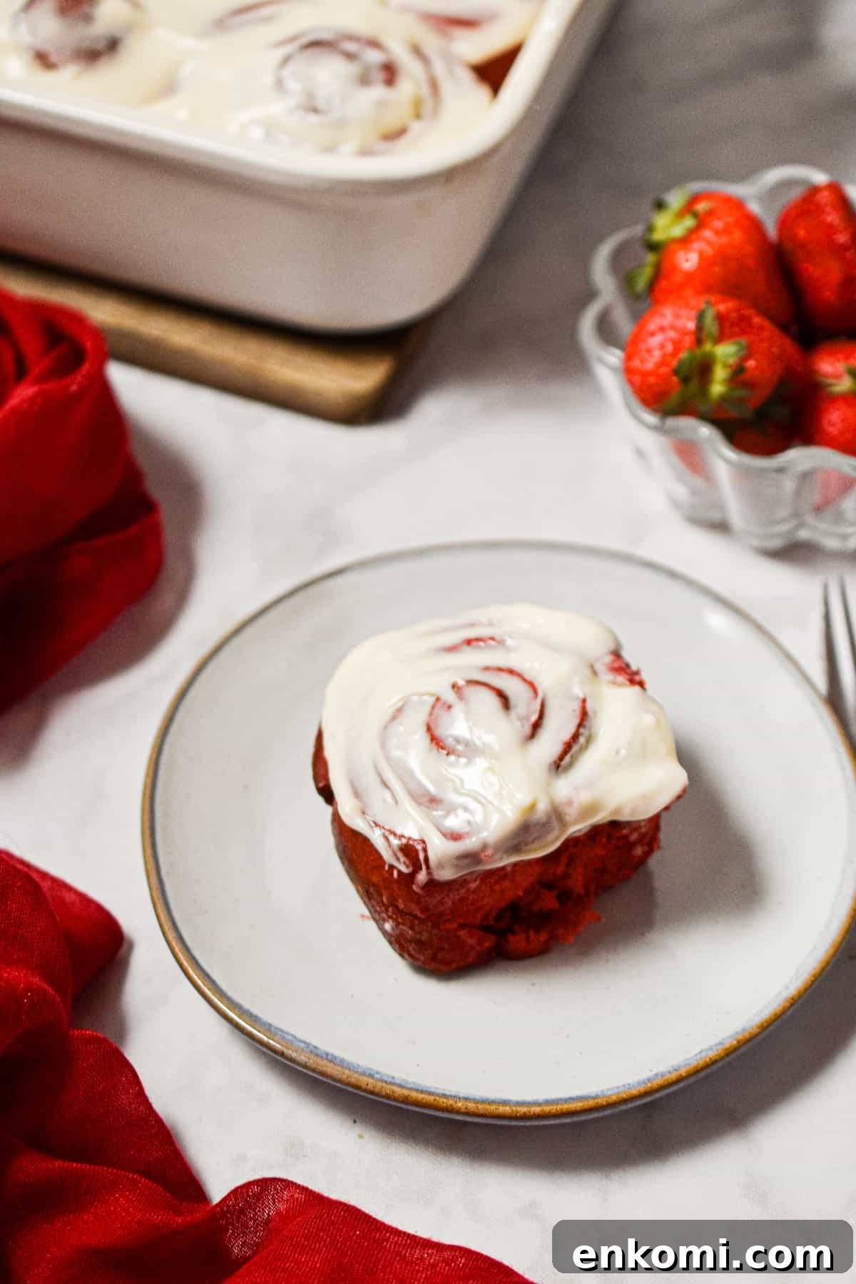 Homemade Red Velvet Cinnamon Rolls with Cream Cheese Glaze 9 One red velvet cinnamon roll is on a plate with a bowl of strawberries next to the baking dish.