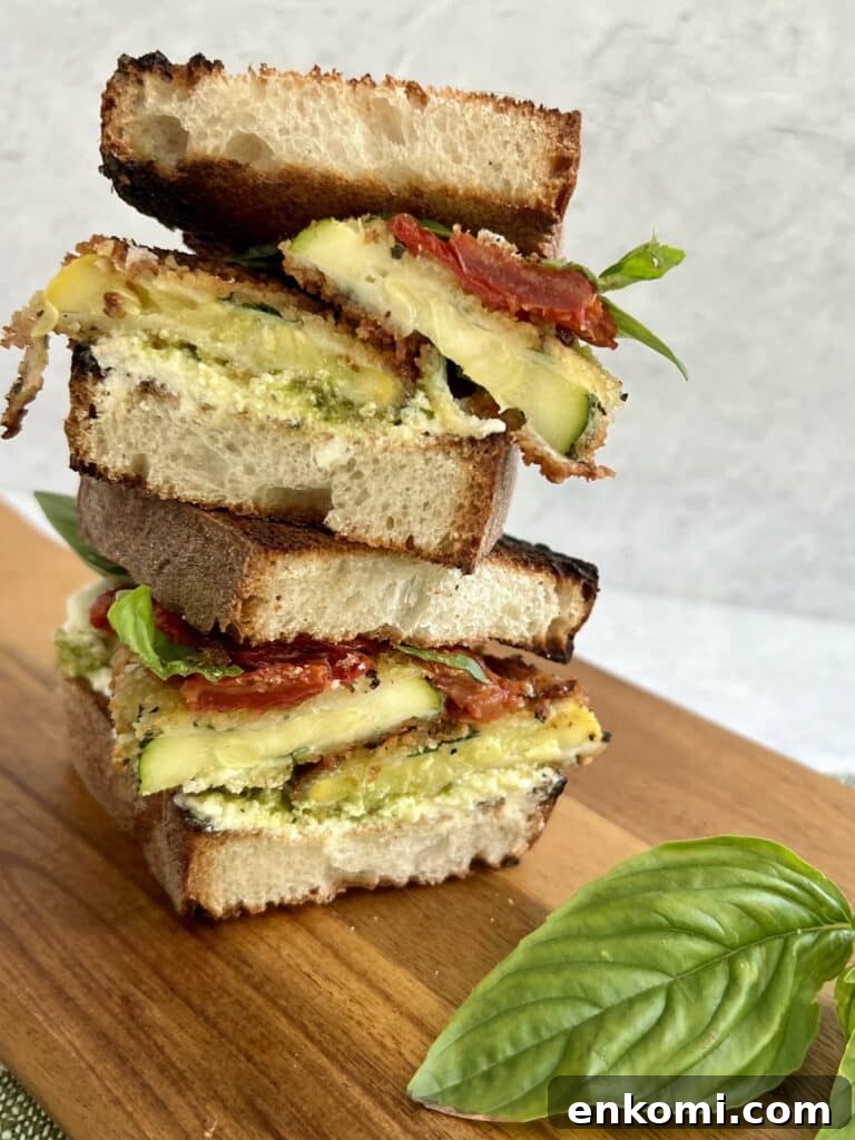 Two halves of a sandwich made with baked panko crusted zucchini, pesto, ricotta and roasted tomatoes stacked on a cutting board with basil garnish.