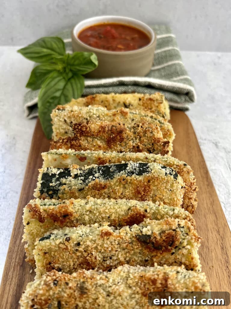 Baked panko crusted zucchini shingled on wooden board with small bowl of tomato dipping sauce on towel and basil sprig garnish.
