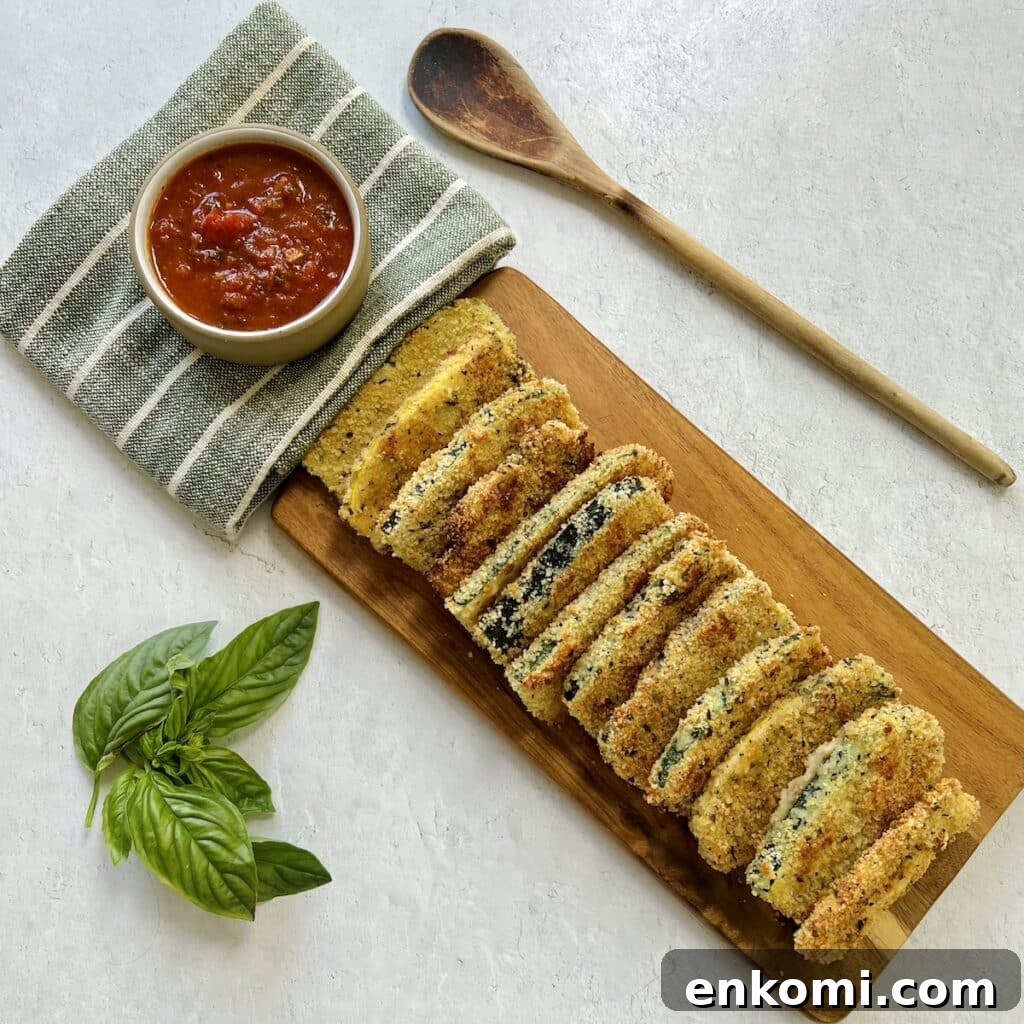 Panko crusted baked zucchini arranged and shingled on wooden board with small bowl of tomato dipping sauce on towel and wooden spoon and basil sprig nearby.