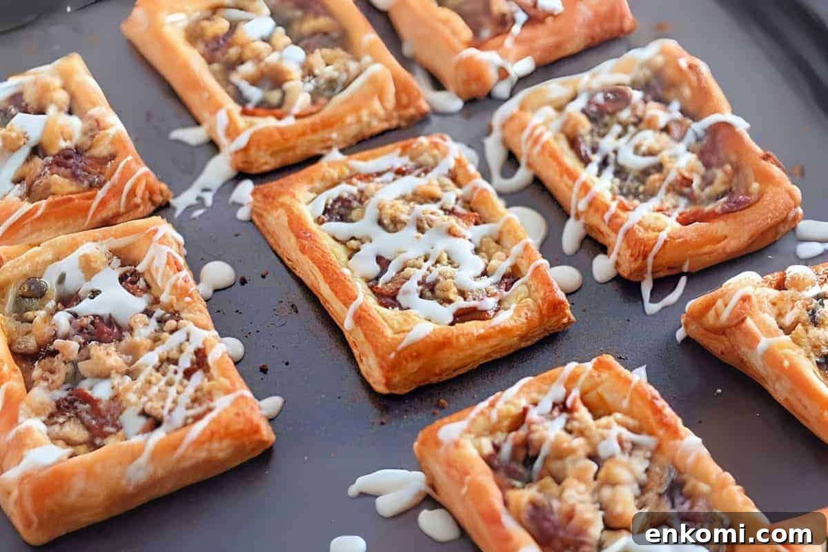 29 Fall Desserts That Bring Generations of Flavor Together 10 Golden brown Dutch Apple Danishes, topped with a fruit and nut filling, drizzled with white icing, displayed on a dark baking sheet.