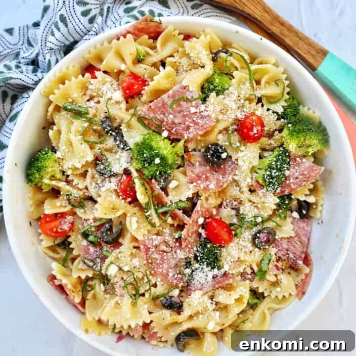 Italian bowtie pasta salad with salami, olives, tomatoes, broccoli, and parmesan cheese in a white bowl with title text that reads "Italian Bow Tie Pasta Salad."