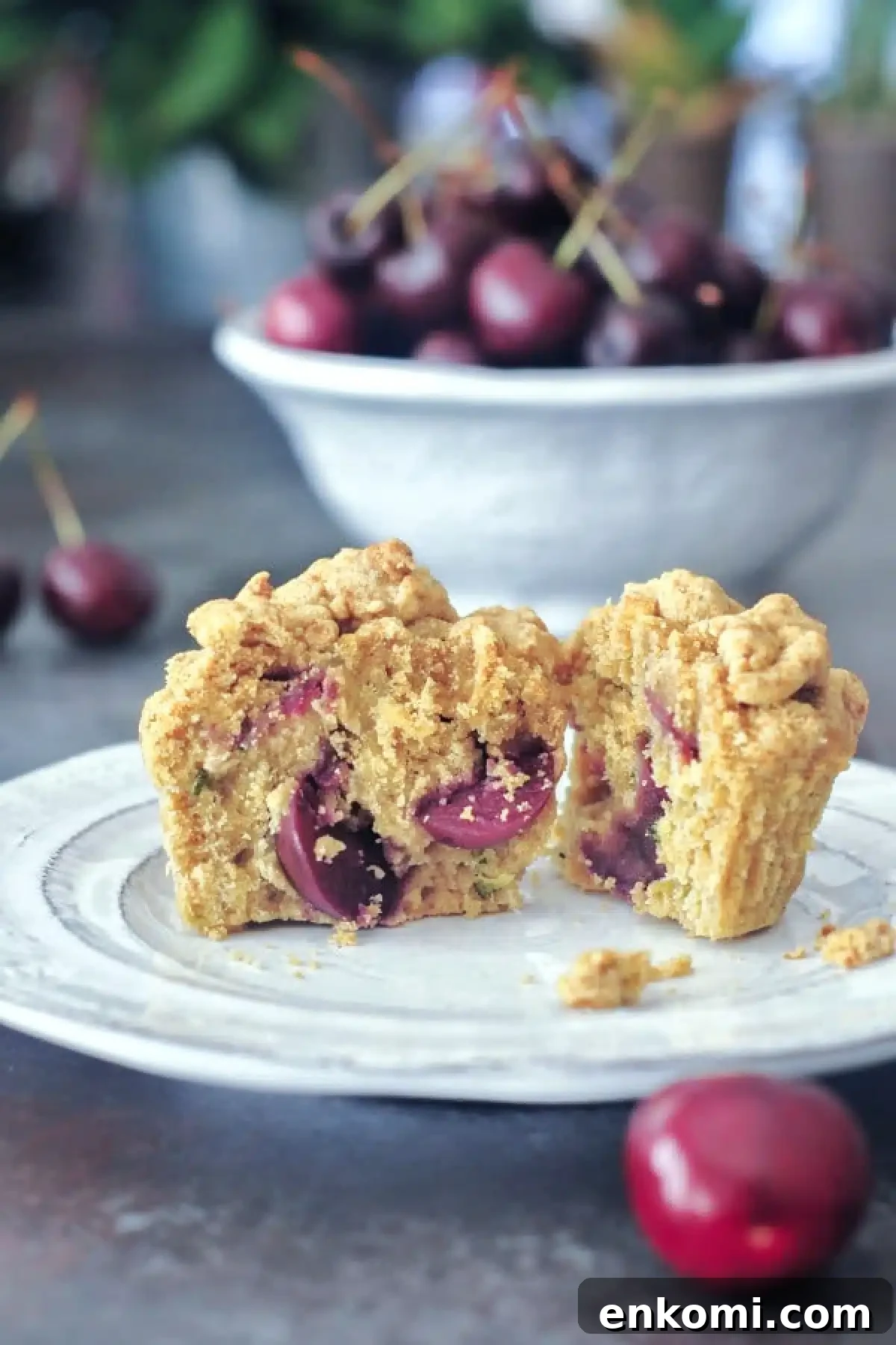 Cherry zucchini muffin sliced in half sitting on a white plate, with a bowl of cherries in background.