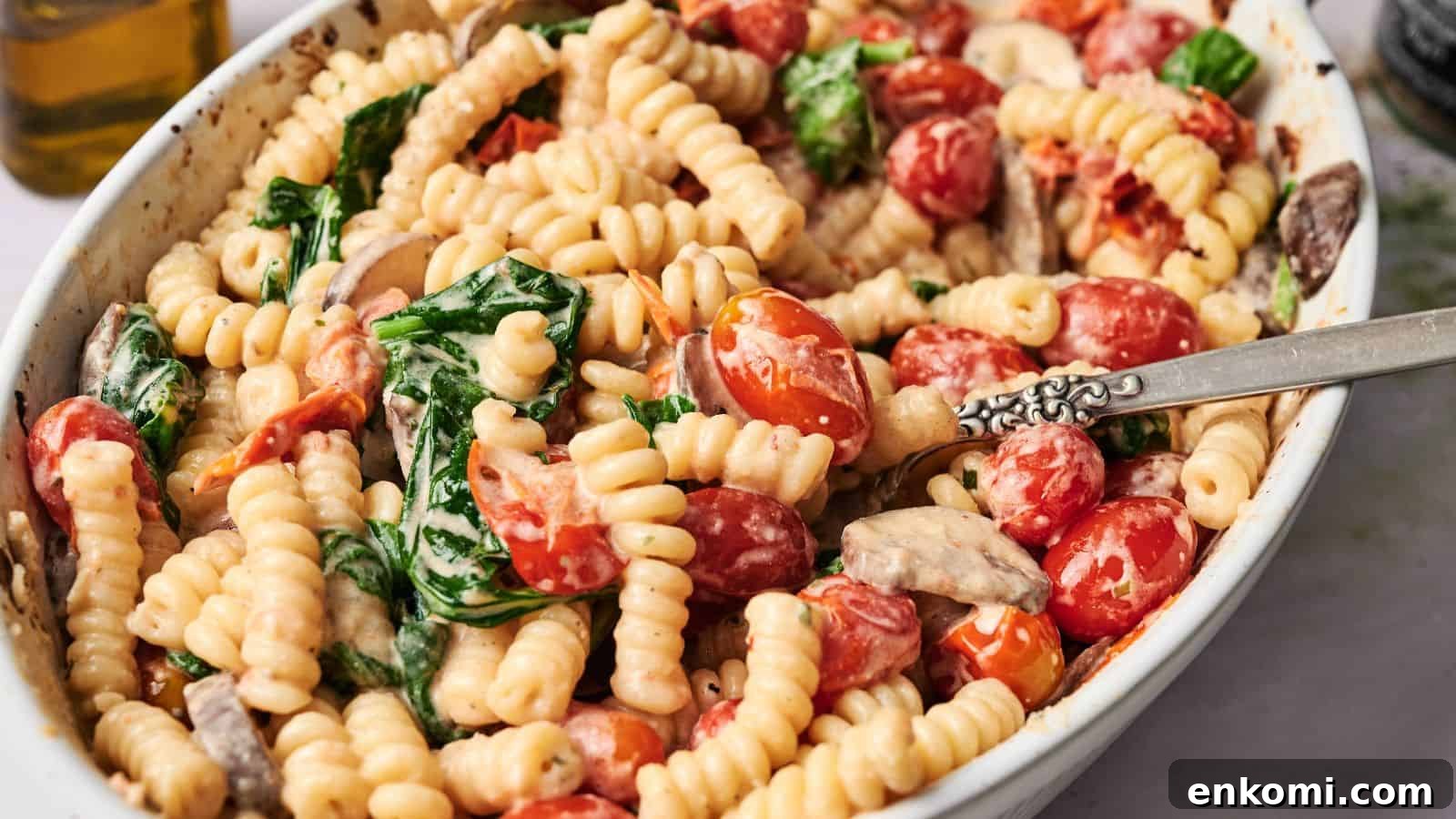 31 Spring Dinners That Feel Fresh, Not Fussy 12 A white dish filled with cooked fusilli pasta, cherry tomatoes, spinach, and mushrooms in a creamy sauce, with a silver fork resting inside.