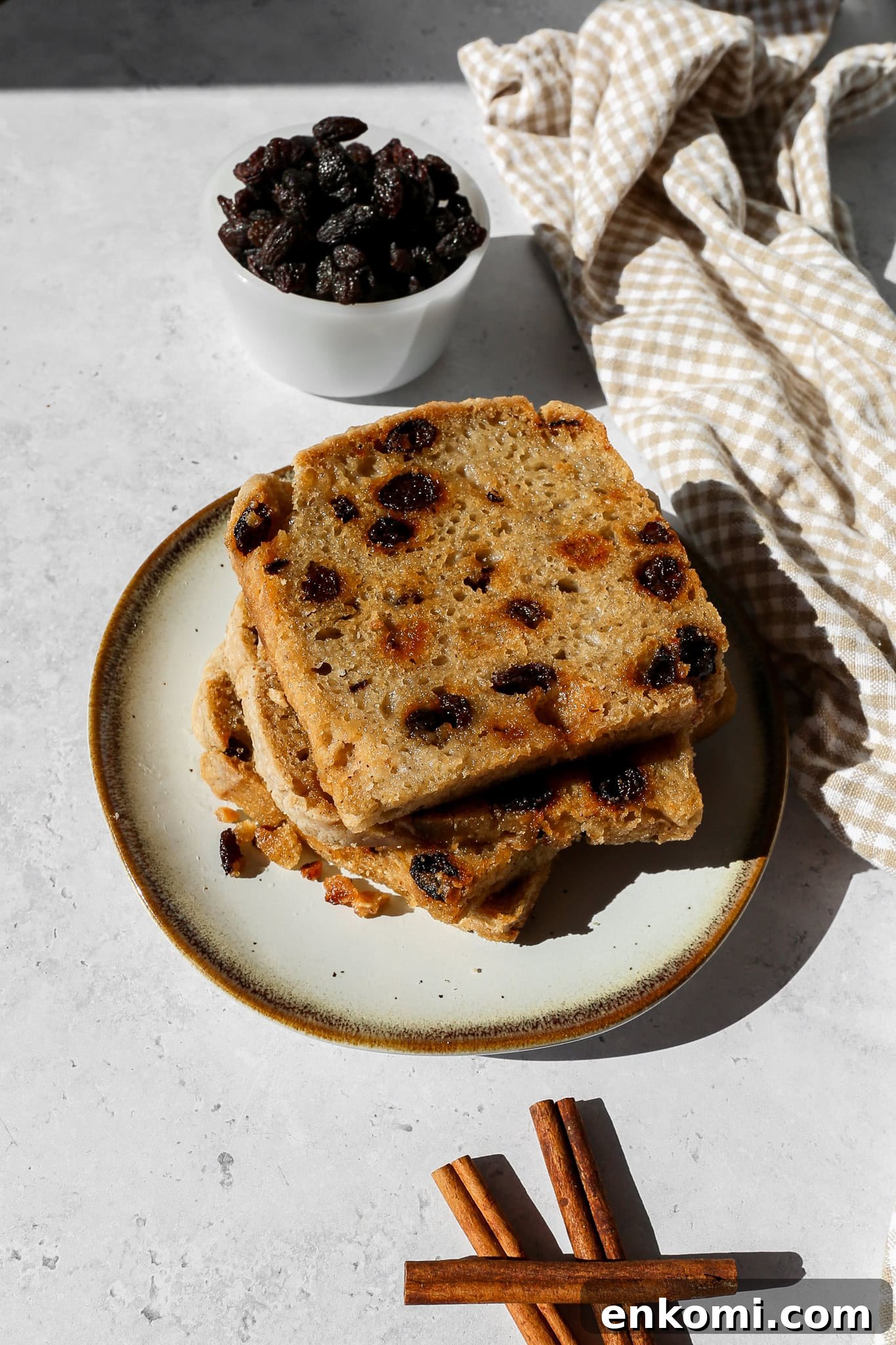 A plate with three slices of toasted gluten free cinnamon raisin sourdough bread next to a small bowl of raisins, cinnamon sticks and a tan and white gingham kitchen towel