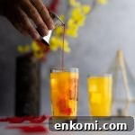 Grenadine being poured into an amaretto sunrise cocktail, with rose petals scattered around and red and yellow flowers in the background.