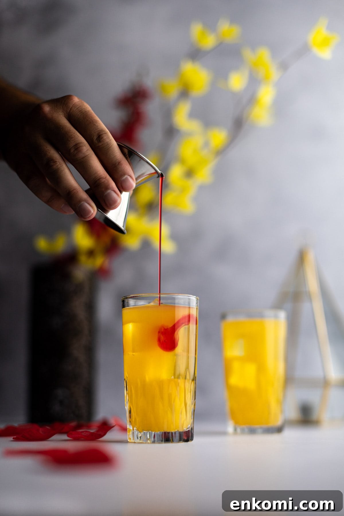 Amaretto Sunrise Cocktail Recipe with Orange and Cherry 3 Hand holding a metal jigger with grenadine syrup being poured into an amaretto cocktail.