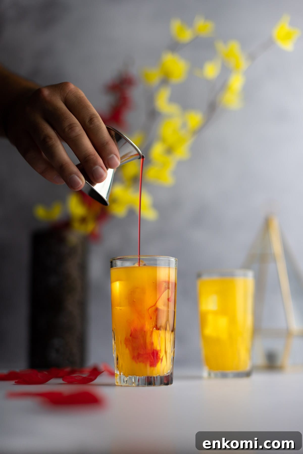 Amaretto Sunrise Cocktail Recipe with Orange and Cherry 4 Hand holding grenadine in a shot glass, pouring grenadine into a cocktail, with grenadine dispersing into the cocktail.