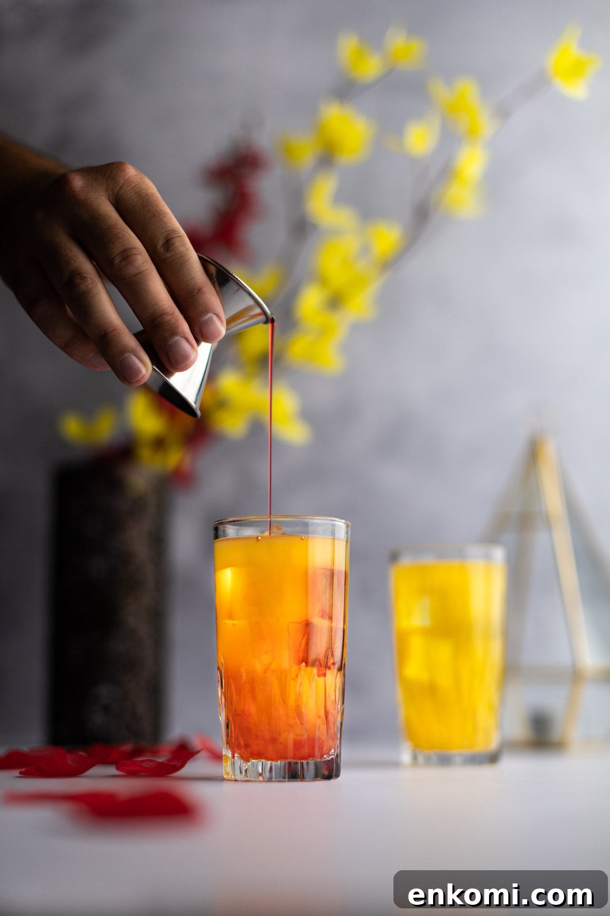 Amaretto Sunrise Cocktail Recipe with Orange and Cherry 5 Grenadine syrup being poured into a yellow cocktail, half mixed with the peach nectar, red and yellow flowers in the background.