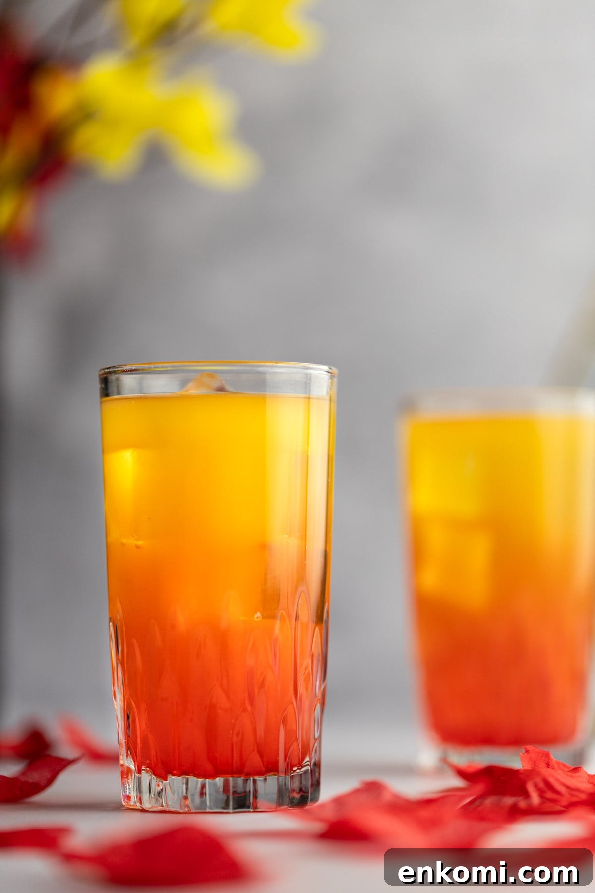 Amaretto Sunrise Cocktail Recipe with Orange and Cherry 6 Close up of an amaretto sunrise cocktail, with rose petals scattered around the drink and flowers in the background.