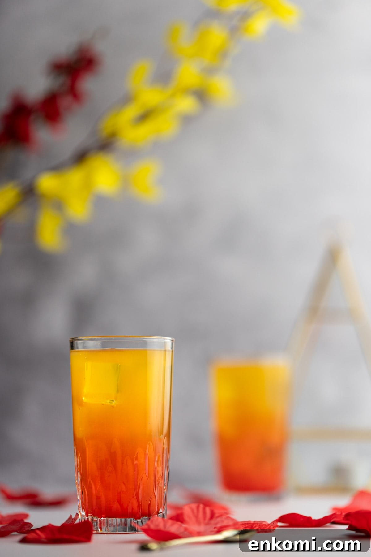 Amaretto Sunrise Cocktail Recipe with Orange and Cherry 7 Two sunrise cocktails, with rose petals scattered around them and flowers in the background.