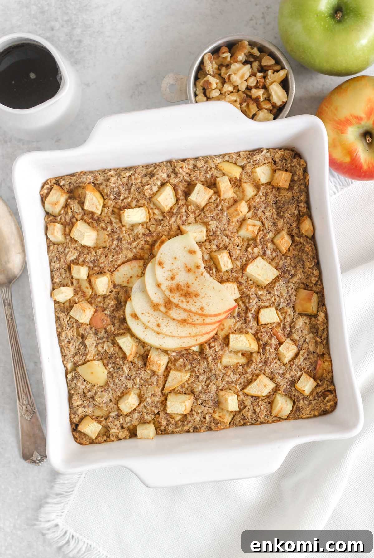 Warm Apple Cinnamon Baked Oatmeal with Crunchy Topping 2 Apple cinnamon baked oatmeal topped with apple sliced and cinnamon in a white square baking dish with apples and syrup off to the side.