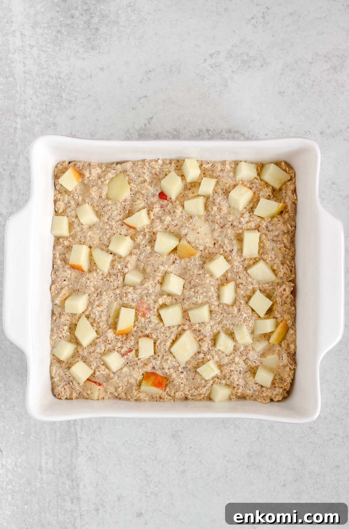 Warm Apple Cinnamon Baked Oatmeal with Crunchy Topping 6 Oatmeal batter in a square white baking dish sprinkled with apple pieces.
