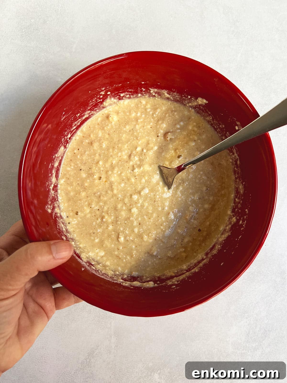 Cheese latke batter mixed in a red bowl with a hand holding it on the left and a fork leaning on the right.