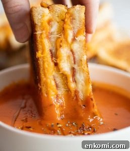 close up shot of hand dunking grilled cheese half into small white bowl of tomato soup