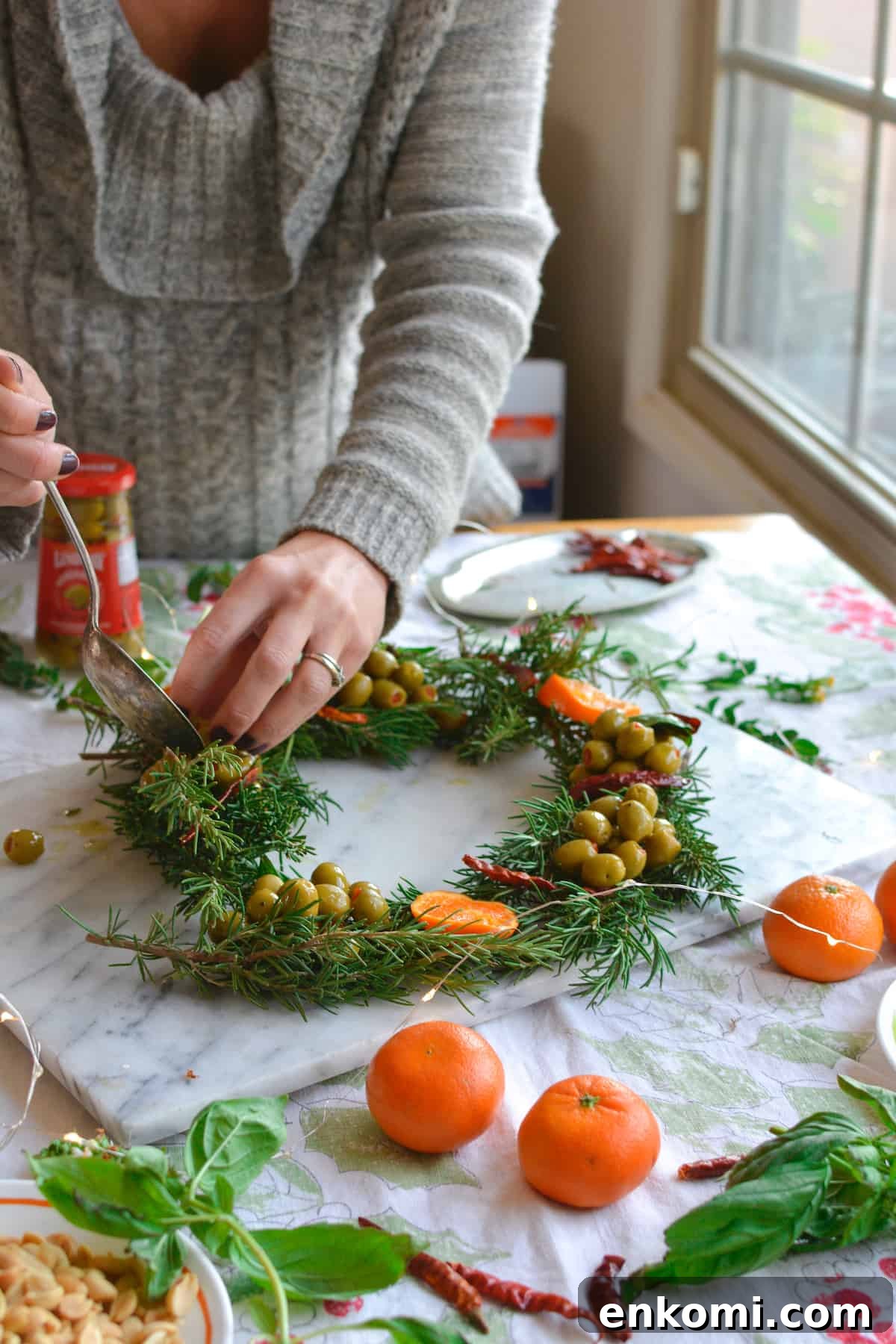 Woman arranging rosemary sprigs and olives on a marble platter to create a wreath shaped appetizer.
