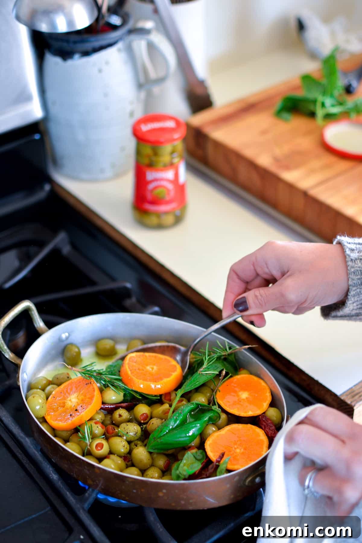 A person warming green, pimento-stuffed olives on the stove with tangerine slices, and fresh herbs in a copper pan.