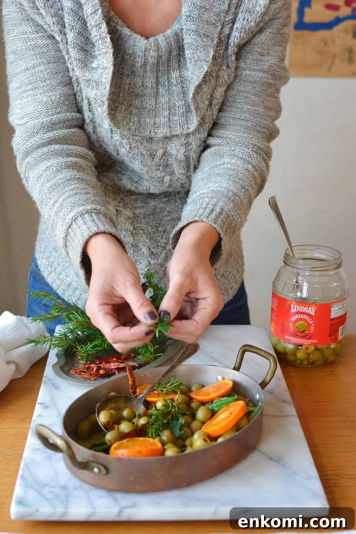 A woman picking the leaves off fresh herbs and putting them into a pan of warmed olives with tangerine slices sitting on a marble platter.