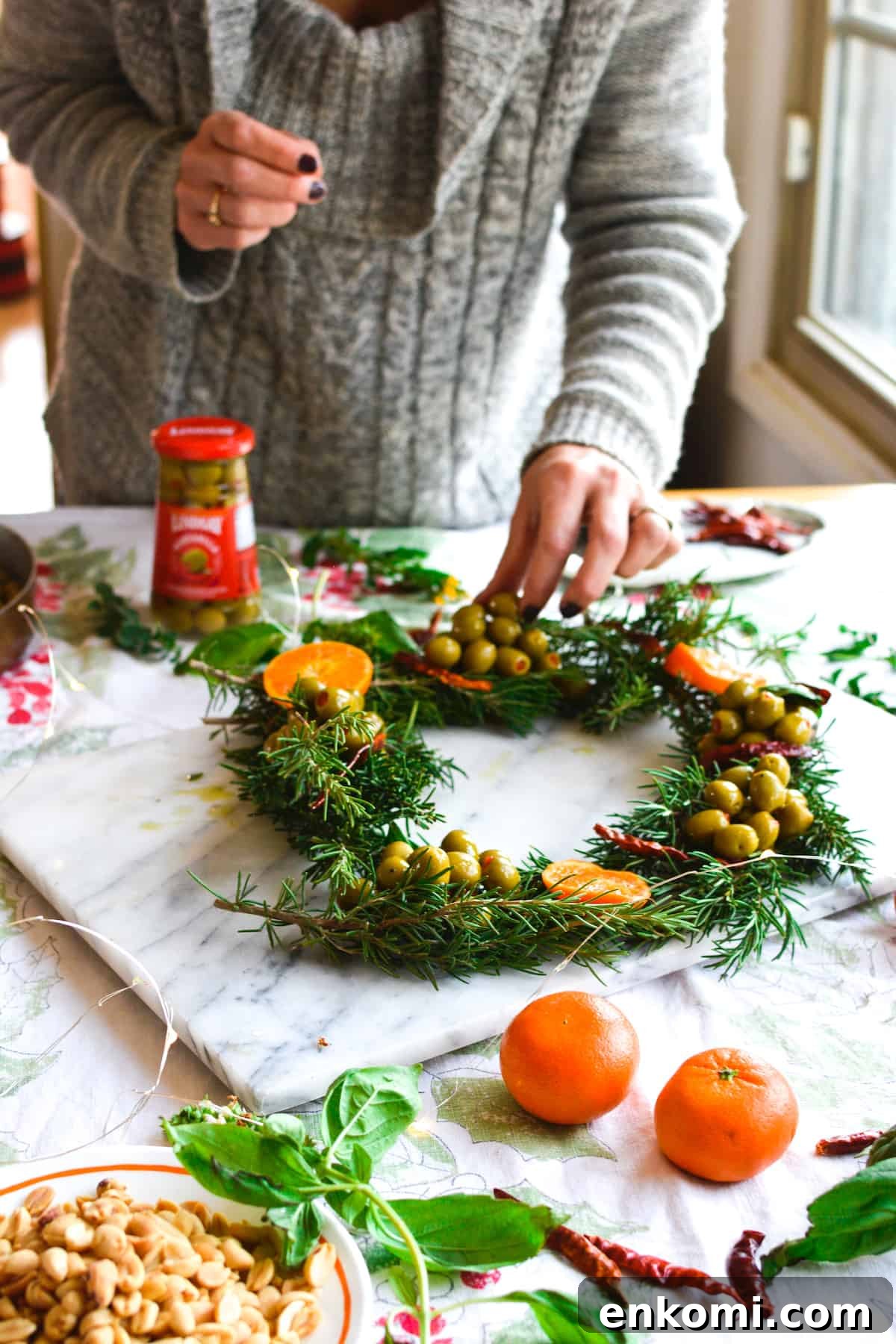 Woman arranging rosemary sprigs and olives on a marble platter to create a wreath shaped appetizer.