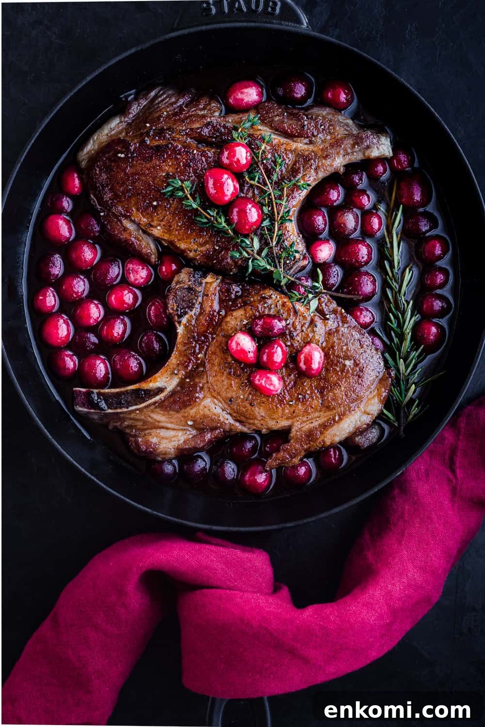 Post sear and pre-oven pork chop overhead shot; chops are with the cranberry sauce in a cast iron skillet with a red linen on the handle; black background.