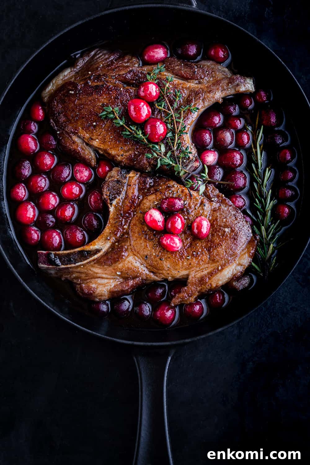Post sear and pre-oven pork chop overhead shot; chops are in a cast iron skillet with the cranberry sauce on a black background.